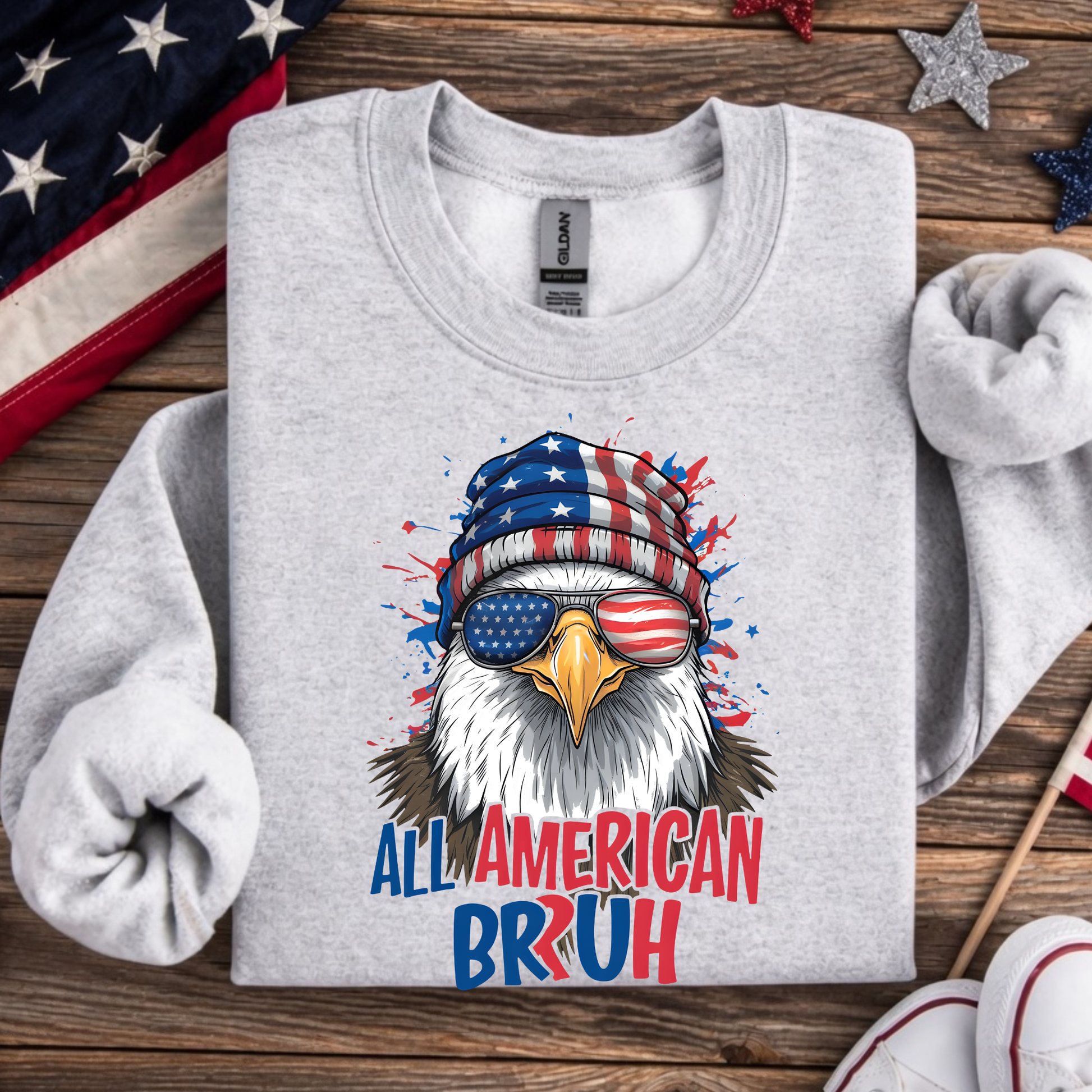 Gray sweatshirt with an eagle wearing a USA flag beanie and sunglasses, text 'All American Bruh' on a wooden surface with American flags.