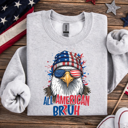 Gray sweatshirt with an eagle wearing a USA flag beanie and sunglasses, text 'All American Bruh' on a wooden surface with American flags.