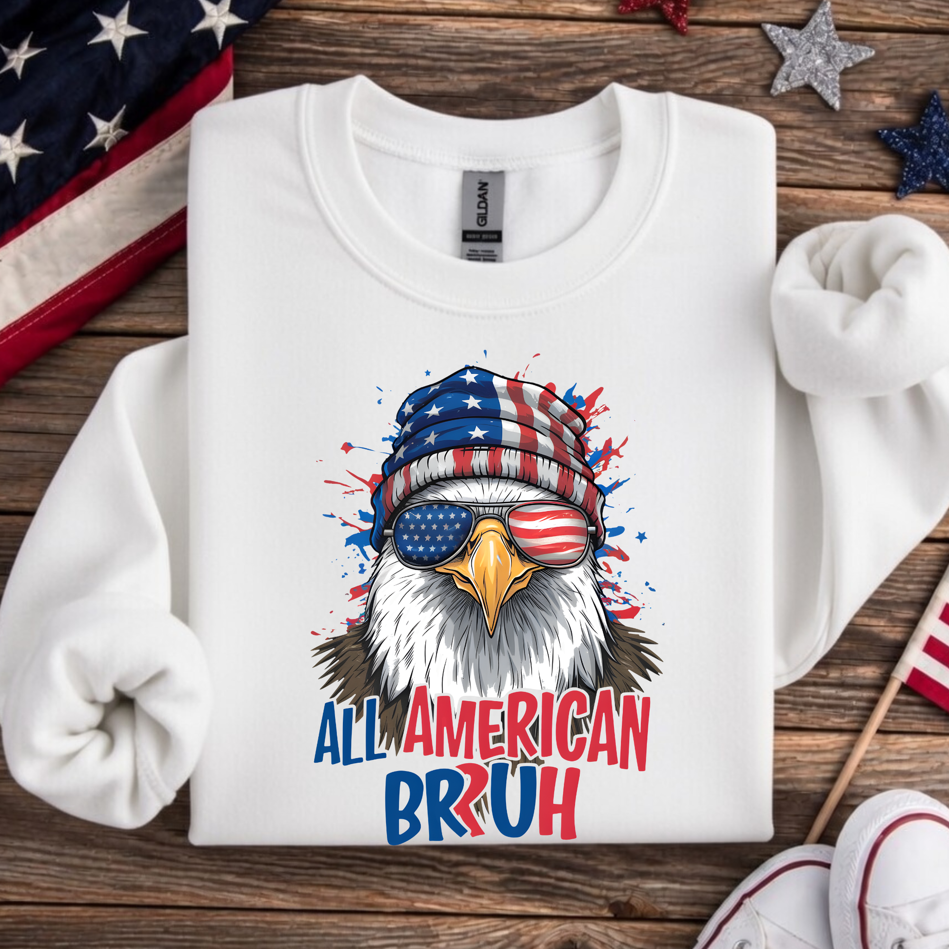 White sweatshirt with an eagle graphic wearing an American flag beanie and sunglasses, text 'All American Bruh', on a wooden surface with American flags.