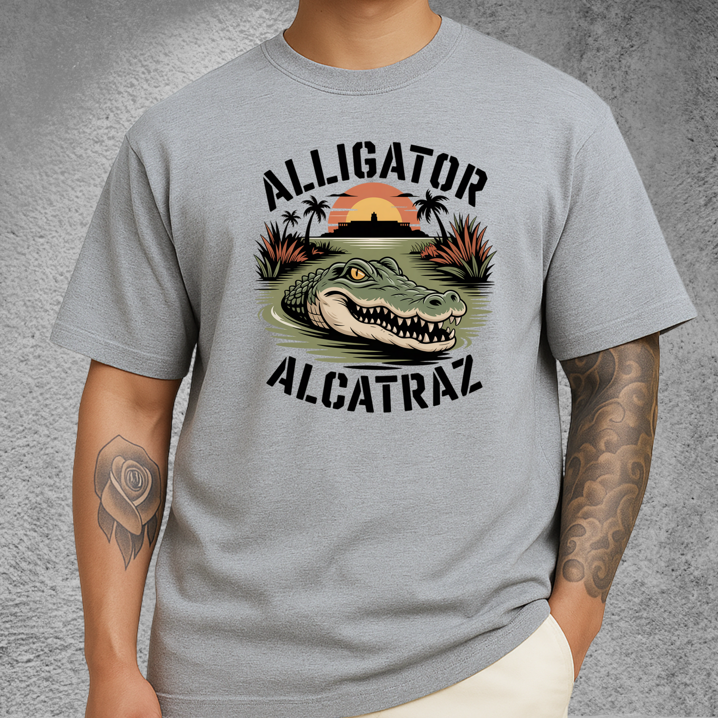 Model wearing a light gray Alligator Alcatraz T-shirt with a fierce gator graphic, Alcatraz silhouette, and sunset background. Unisex men's tee perfect for travelers, nature lovers, and graphic shirt collectors.