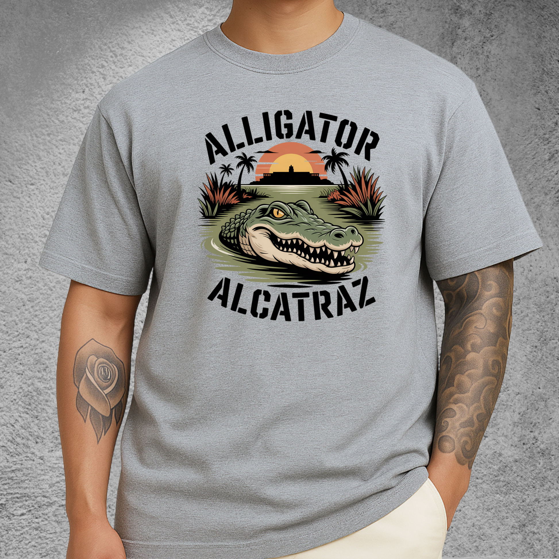Model wearing a light gray Alligator Alcatraz T-shirt with a fierce gator graphic, Alcatraz silhouette, and sunset background. Unisex men's tee perfect for travelers, nature lovers, and graphic shirt collectors.