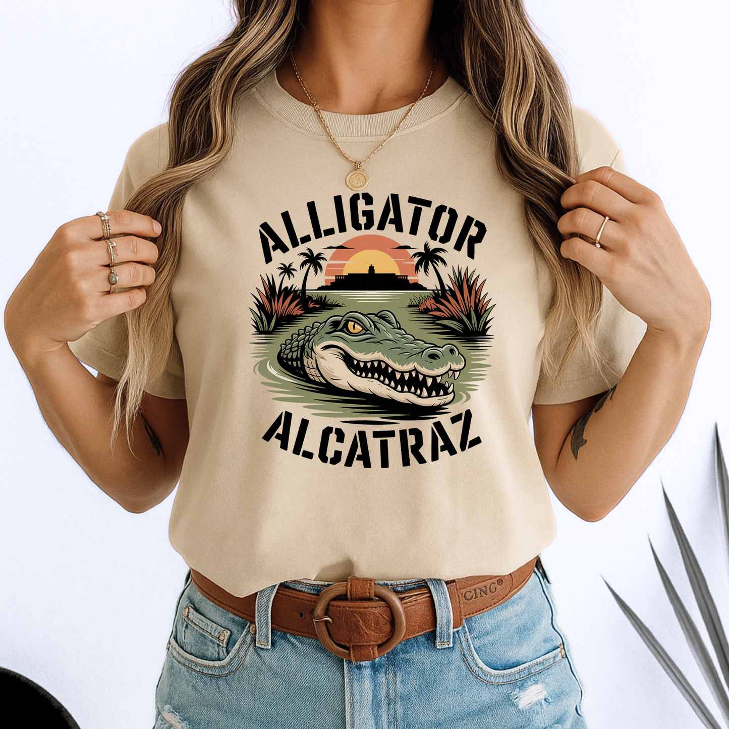 Woman in a beige Alligator Alcatraz T-shirt with vivid gator and sunset design. Soft unisex cotton tee styled with denim, great for travel or casual days.