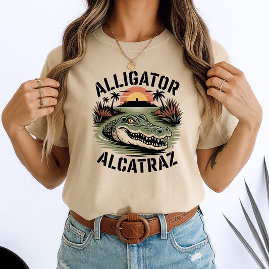 Woman in a beige Alligator Alcatraz T-shirt with vivid gator and sunset design. Soft unisex cotton tee styled with denim, great for travel or casual days.