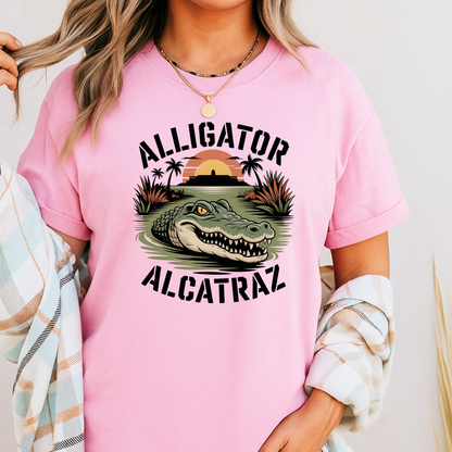 Close-up of a pink Alligator Alcatraz shirt worn by a woman. Features a fierce gator in water with a prison silhouette and palm trees. Playful graphic shirt.
