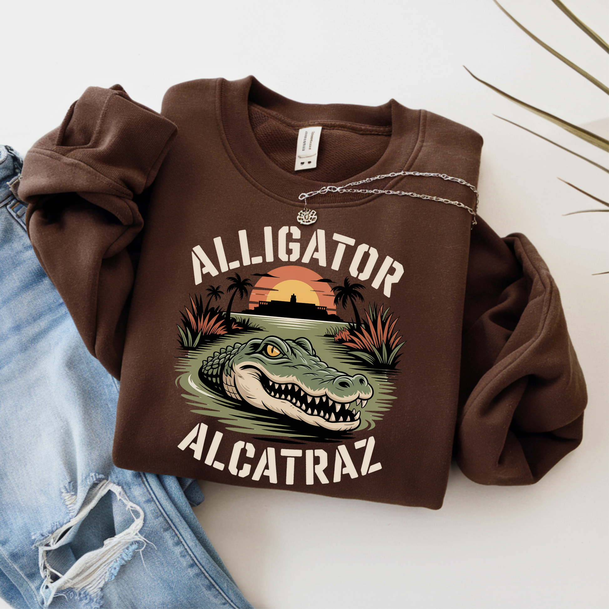 Brown sweatshirt with alligator and Alcatraz design on a white surface with jeans.