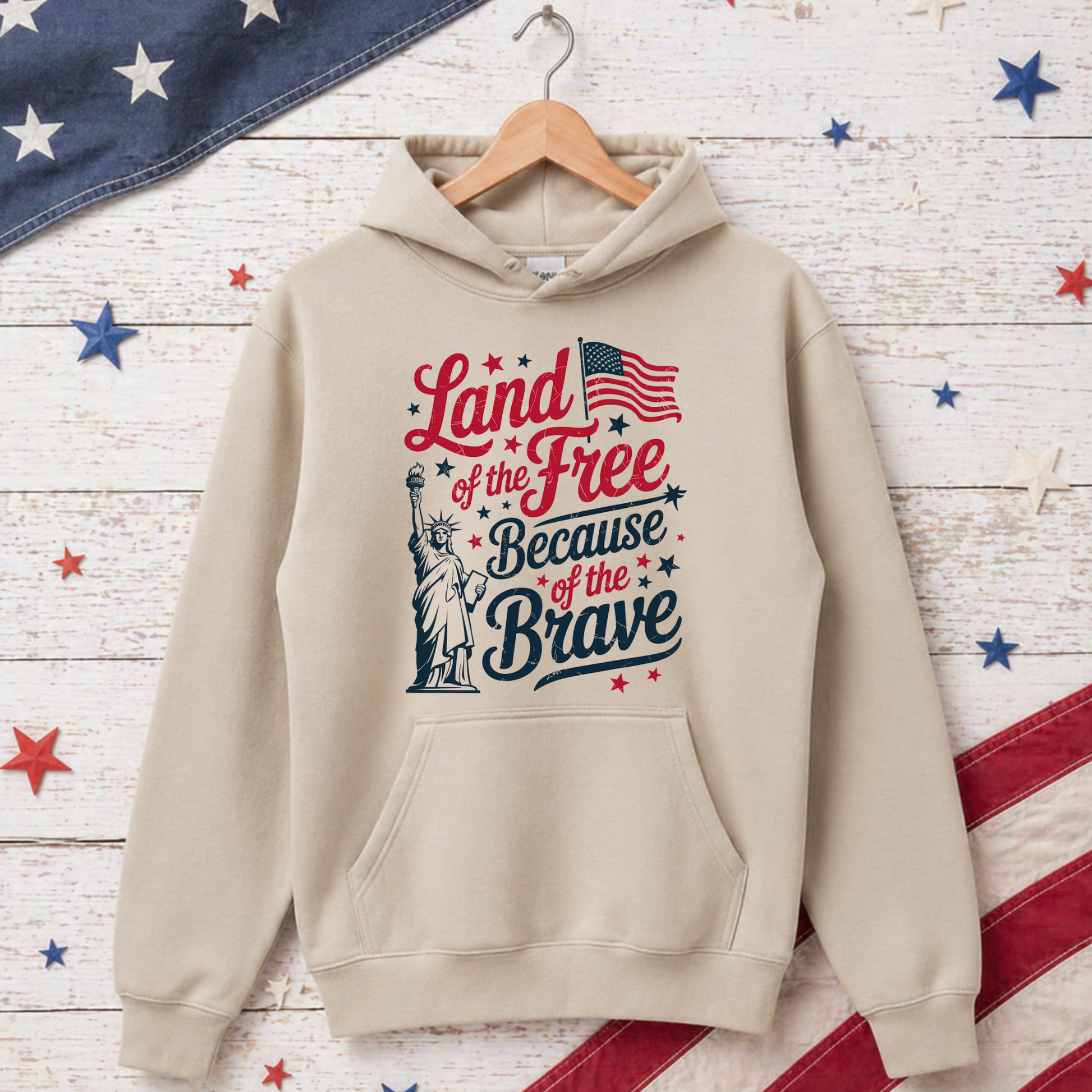 Beige hoodie with 'Land of the Free Because of the Brave' text and Statue of Liberty graphic on a wooden background with American flags.