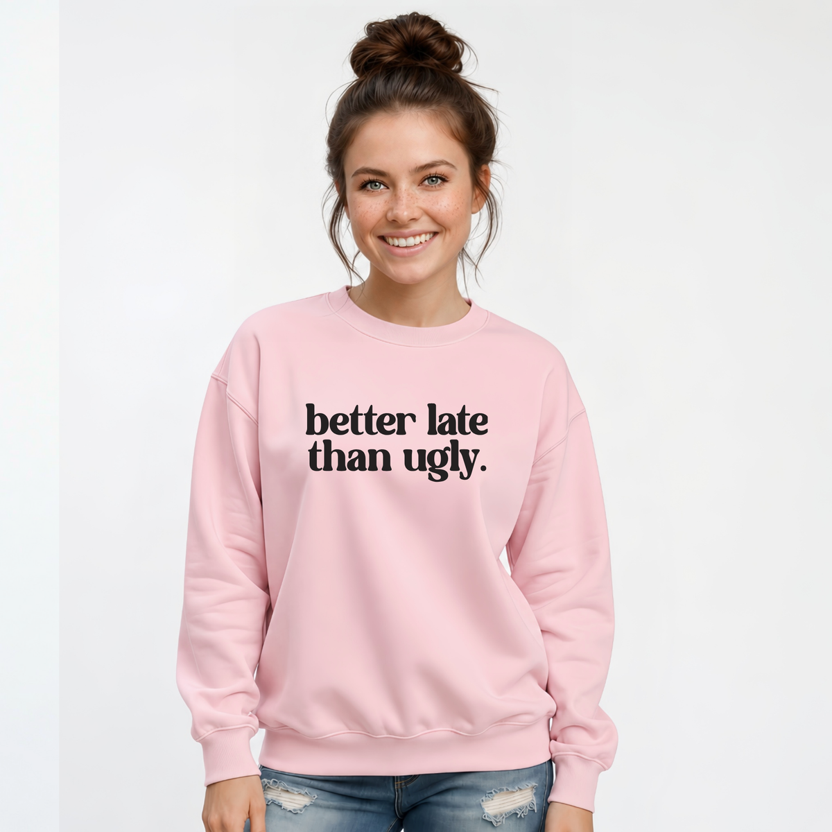 Woman wearing a light pink crewneck sweatshirt with black Better Late Than Ugly lettering against a white studio background.