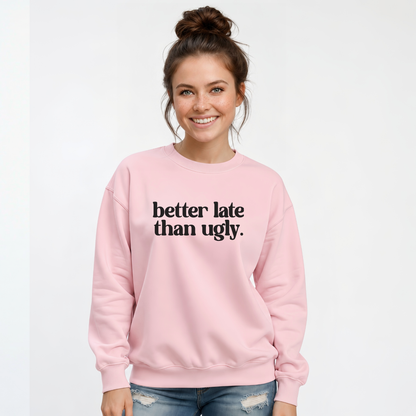 Woman wearing a light pink crewneck sweatshirt with black Better Late Than Ugly lettering against a white studio background.
