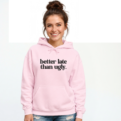 Woman wearing a light pink hoodie with black Better Late Than Ugly lettering against a white studio background.