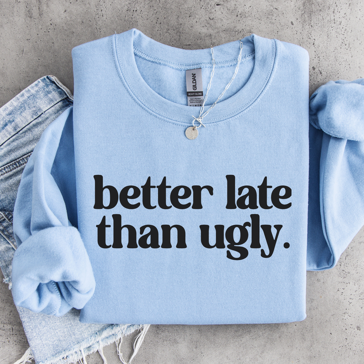 Light blue Better Late Than Ugly crewneck sweatshirt flat lay with black lettering, styled with denim and a necklace.