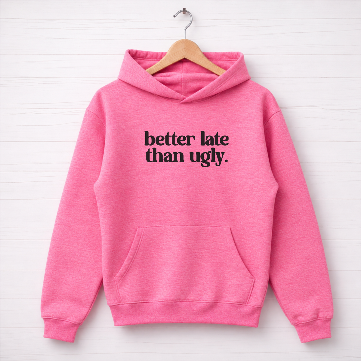 Bright pink Better Late Than Ugly hoodie hanging on a wooden hanger.