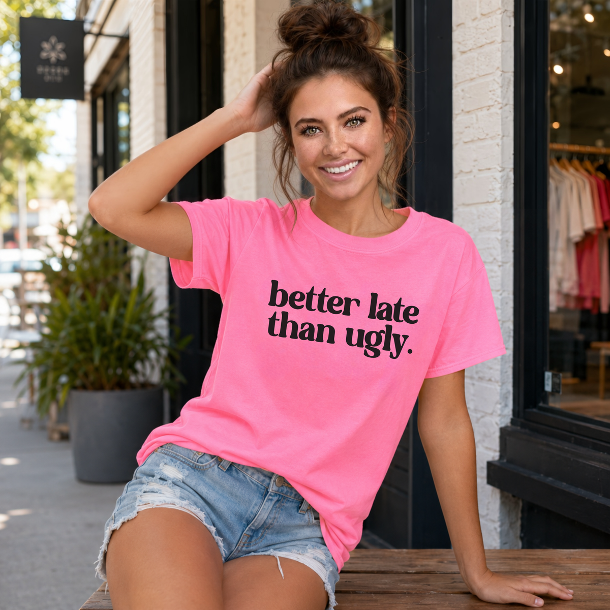 Woman wearing a bright pink Better Late Than Ugly T-shirt while seated outside a modern storefront.