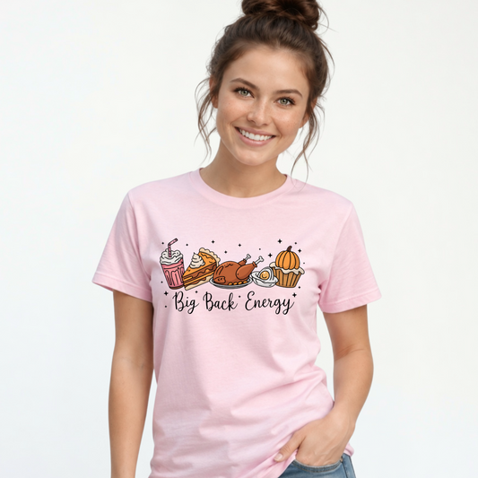 Smiling woman models a light pink T-shirt with a colorful Big Back Energy Thanksgiving food graphic, paired with distressed jeans in a white studio.