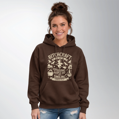 Woman wearing a brown hoodie with a Bitchcraft witch, ravens, cauldron and potion graphic against a light studio background.
