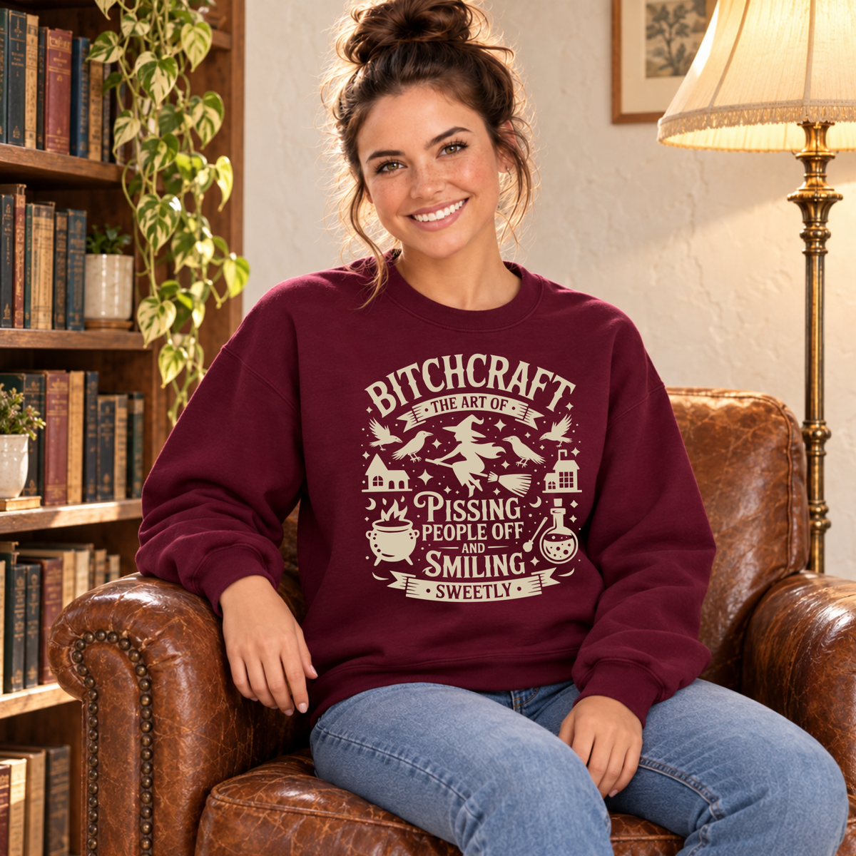 Woman wearing a burgundy crewneck sweatshirt with a Bitchcraft witch, ravens, cauldron and potion graphic while seated in a library.