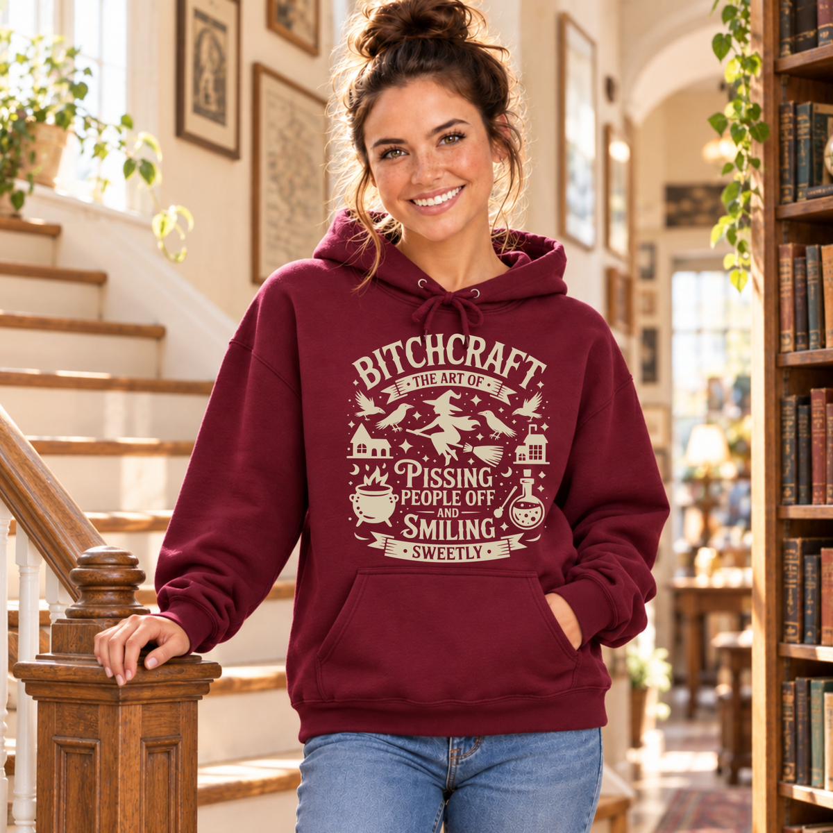 Woman wearing a burgundy hoodie with a Bitchcraft witch, ravens, cauldron and potion graphic beside a staircase and bookshelves.