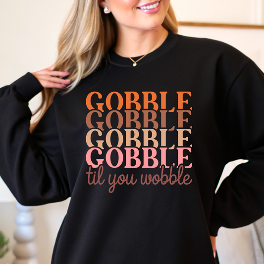 Black cozy fall sweatshirt with “Gobble Gobble” text in orange and pink tones – woman modeling in home setting.