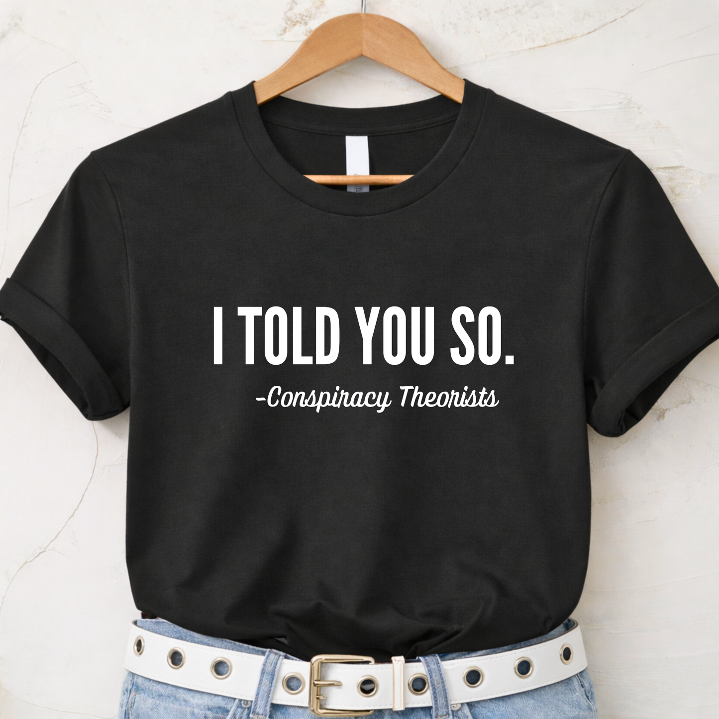 Black t-shirt with 'I Told You So. - Conspiracy Theorists' text on a hanger.