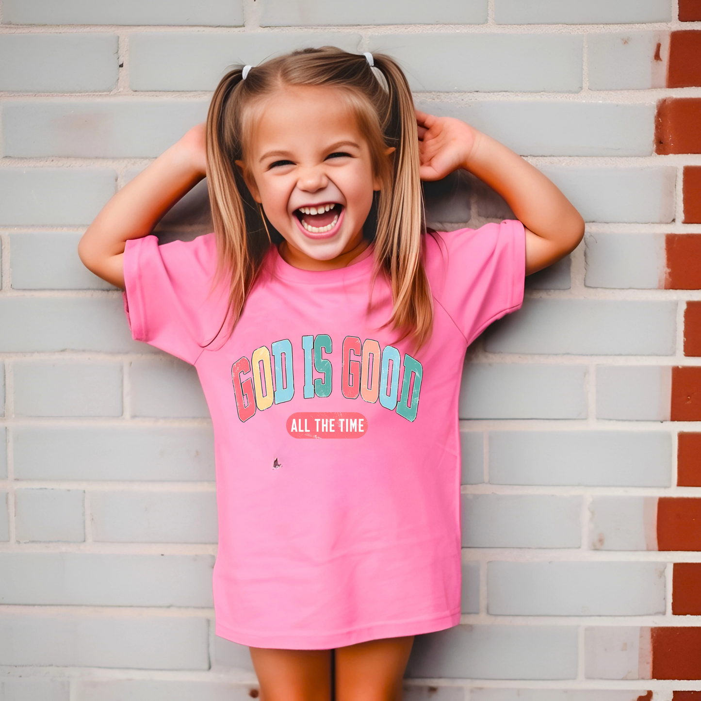 laughing young girl wearing a bright pink T-shirt with the message 'God Is Good All the Time' in colorful text, symbolizing faith and positivity. Perfect for Easter and Christian-themed apparel.