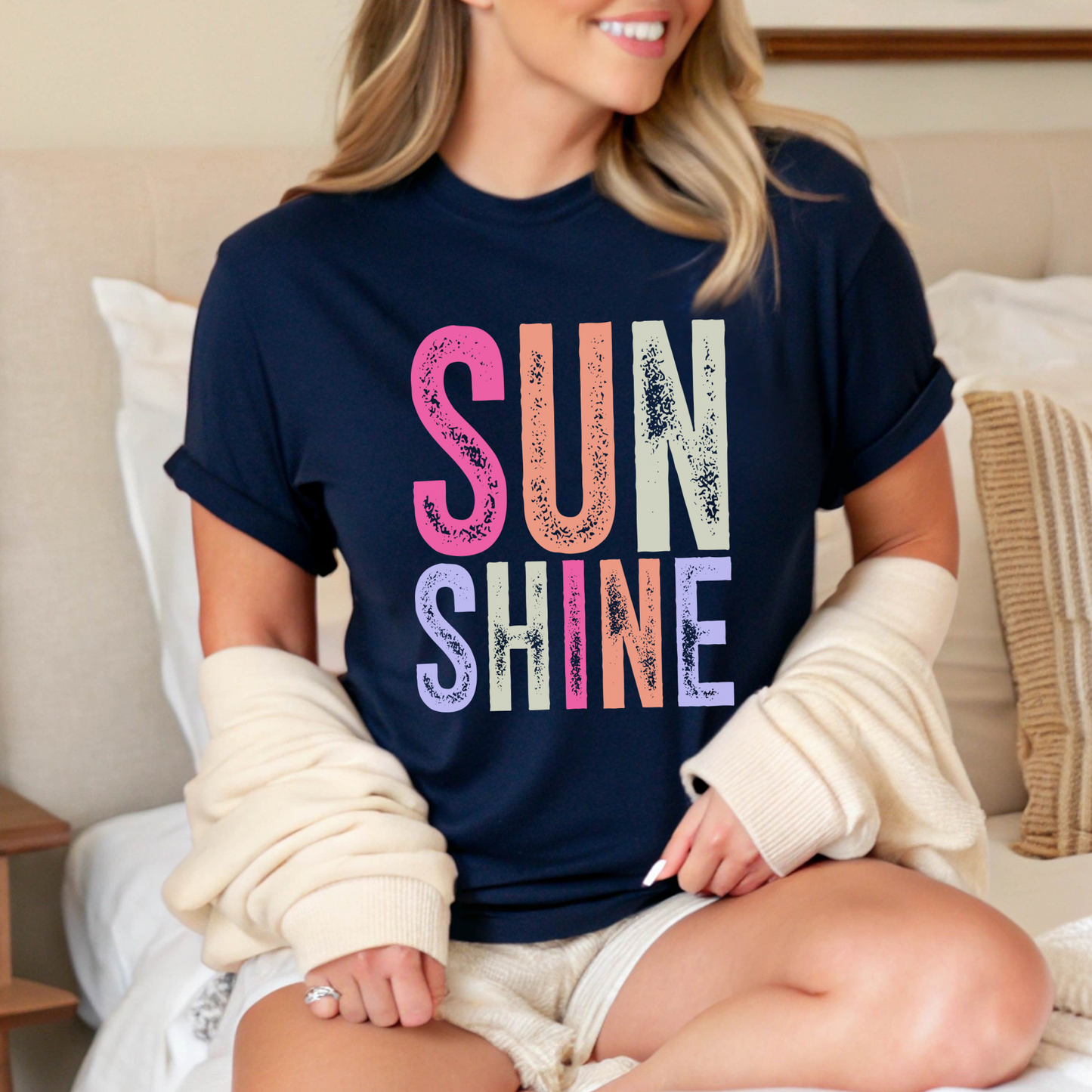 Woman wearing a bright “SUNSHINE” graphic T-shirt in Navy, smiling in a relaxed summer setting.

