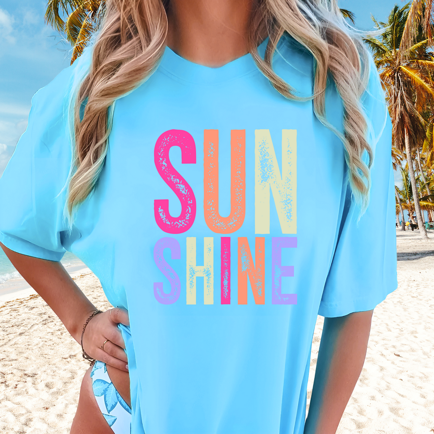 Woman wearing a bright “SUNSHINE” graphic T-shirt in sky blue, smiling in a relaxed summer setting.
