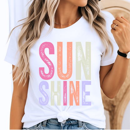 Woman wearing a bright “SUNSHINE” graphic T-shirt in White, smiling in a relaxed summer setting.