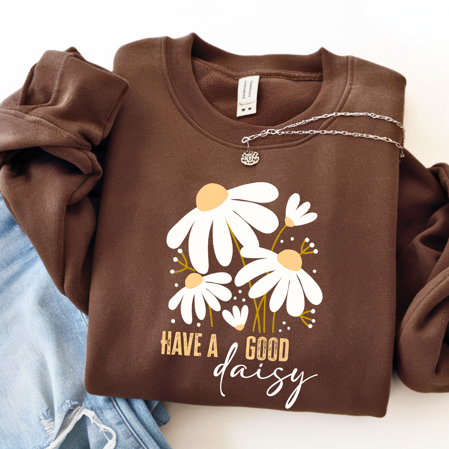 Chocolate brown “Have a Good Daisy” sweatshirt folded with jeans and a necklace, featuring a cozy fall floral graphic perfect for seasonal outfits, pumpkin patch days, or casual autumn layering.
