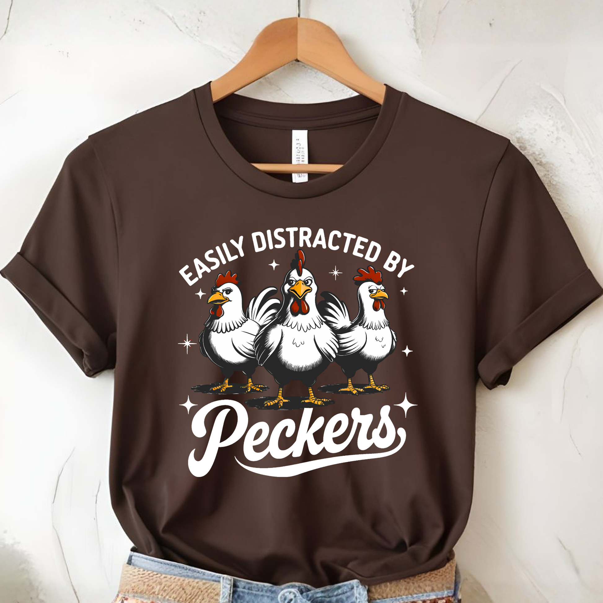 Brown t-shirt with chicken graphic and text on a hanger against a white wall.