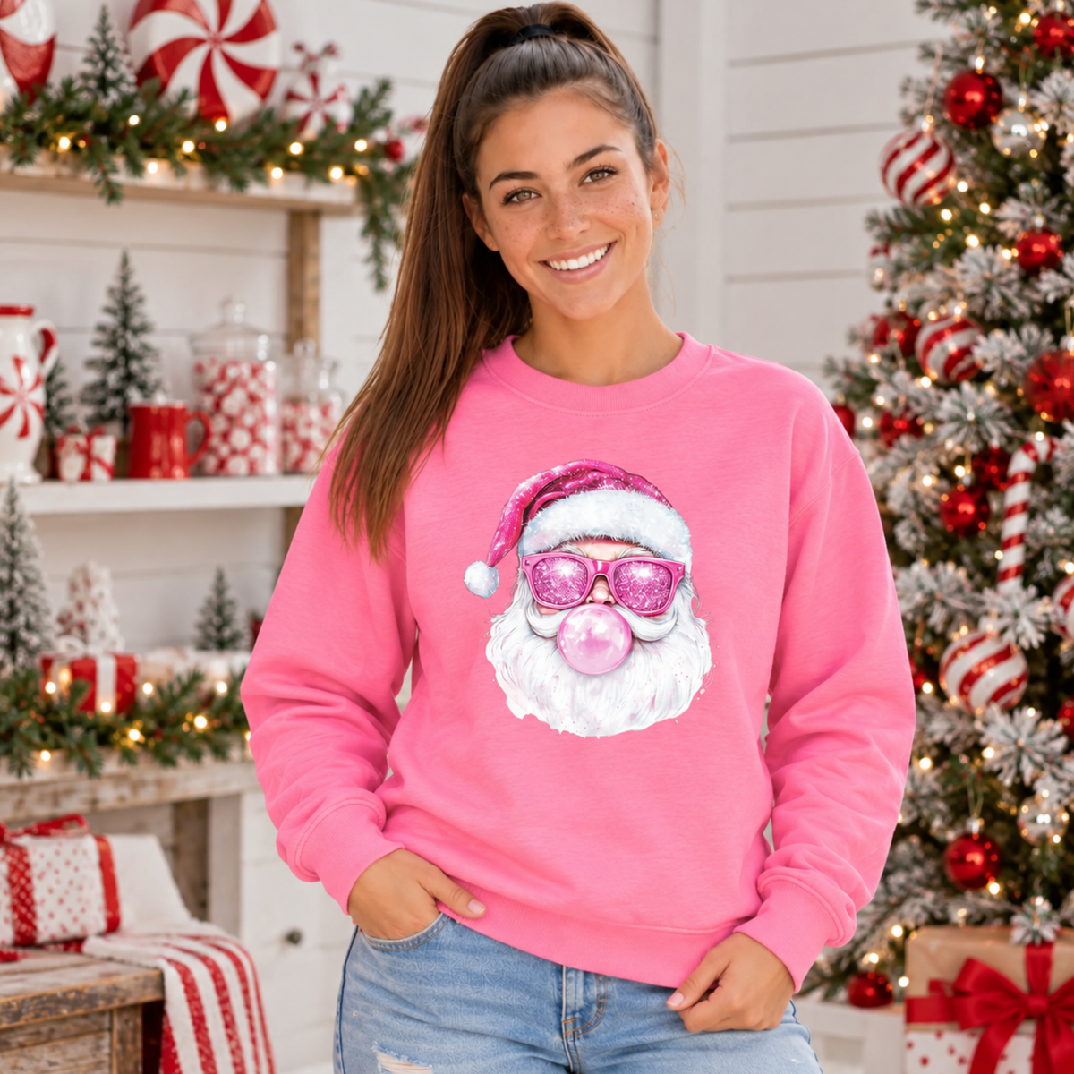 Woman wearing a bright pink Bubblegum Santa crewneck sweatshirt beside a peppermint-decorated Christmas tree