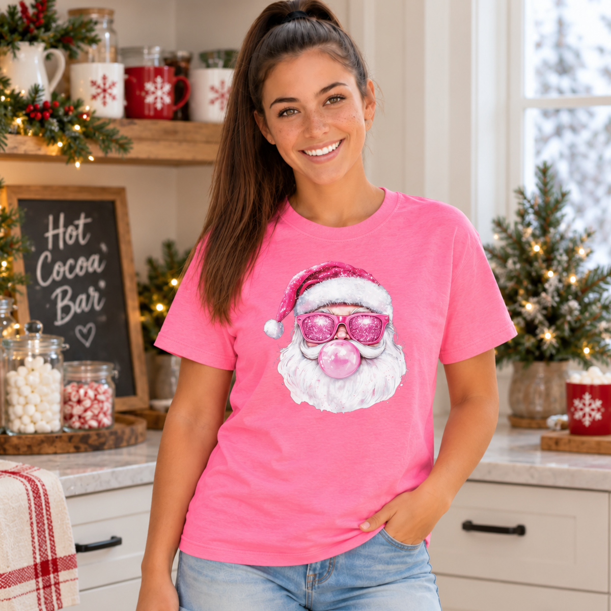 Woman wearing a bright pink Bubblegum Santa T-shirt at a festive hot cocoa bar with candy jars and greenery