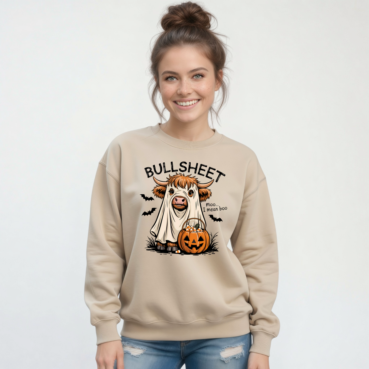 Woman wearing a Sand crewneck sweatshirt with the Bullsheet Halloween Highland cow ghost and jack-o'-lantern design against a white background.