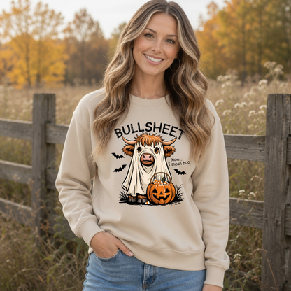 Woman outdoors in a Sand crewneck sweatshirt featuring the Bullsheet Halloween Highland cow ghost and jack-o'-lantern design, with a wooden fence and fall foliage behind her.