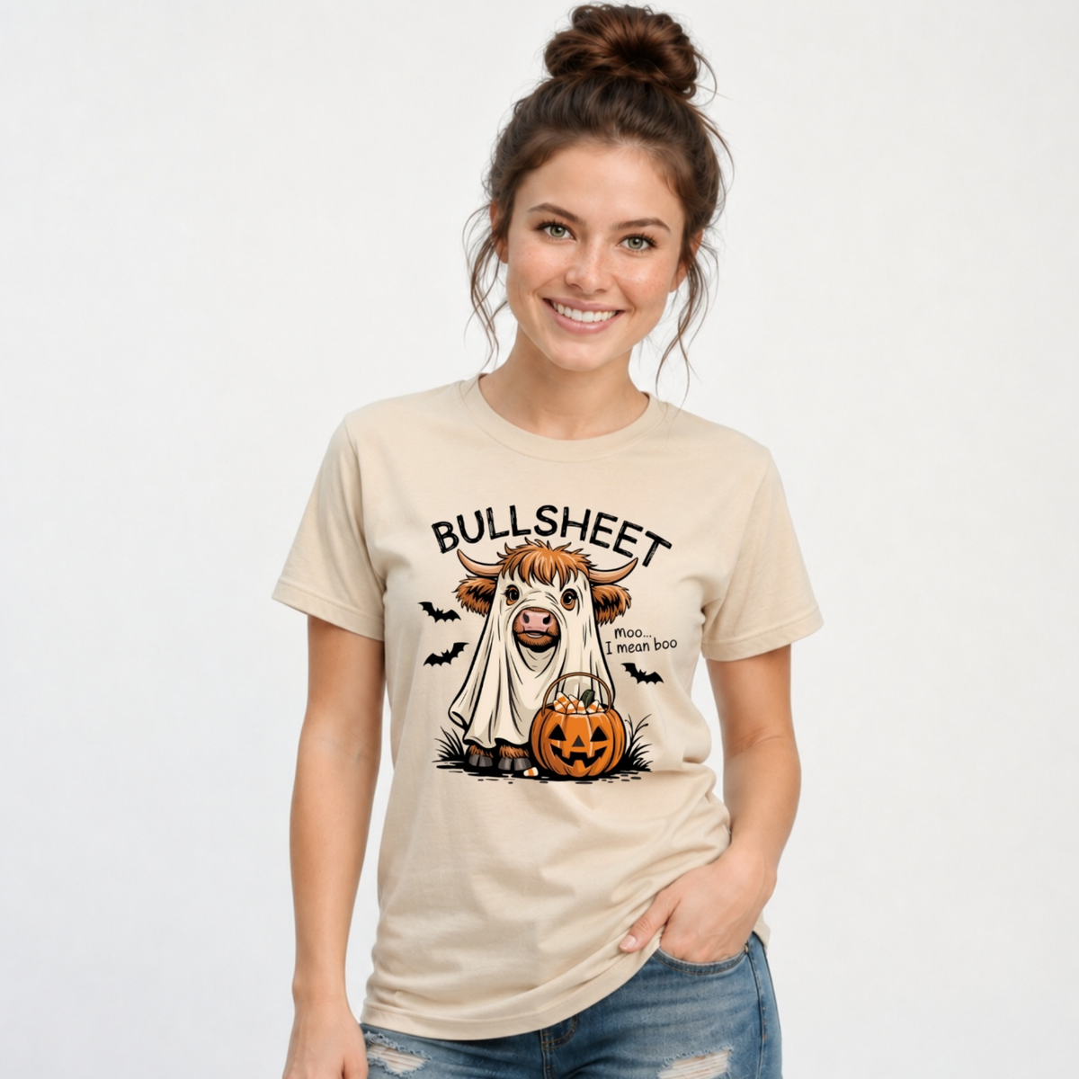 Woman wearing a Sand T-shirt featuring the Bullsheet Halloween Highland cow ghost and jack-o'-lantern design.