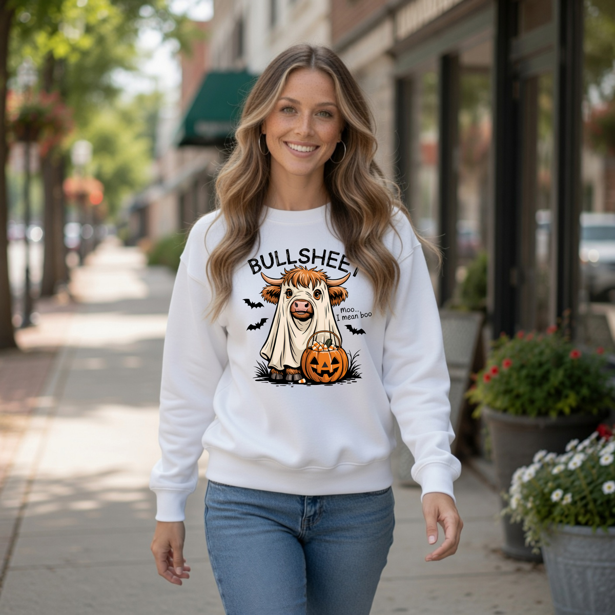 Woman on a city sidewalk wearing a White crewneck sweatshirt with the Bullsheet Halloween Highland cow ghost and jack-o'-lantern design.