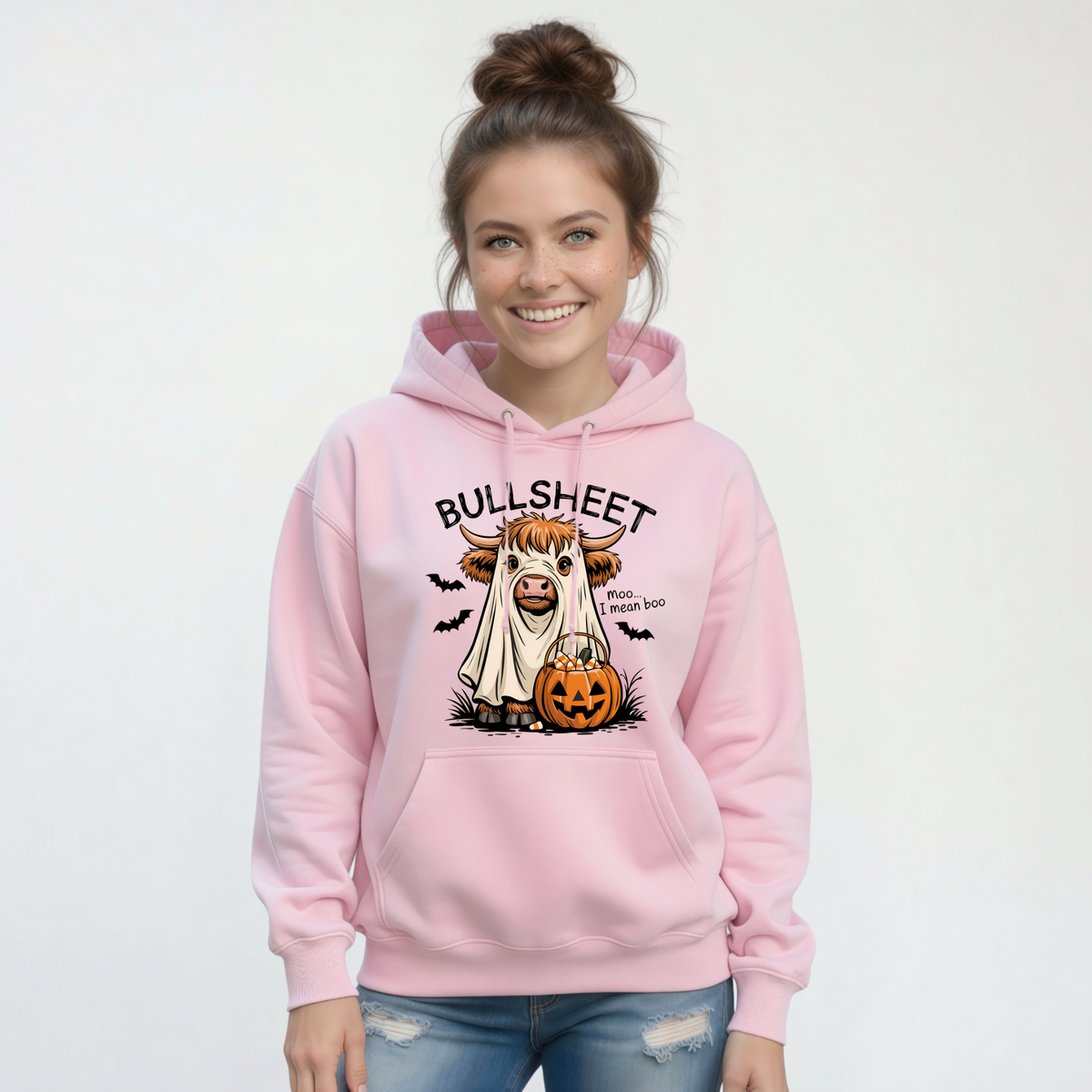 Smiling woman wearing a Light Pink pullover hoodie featuring the Bullsheet Halloween Highland cow ghost and jack-o'-lantern design.