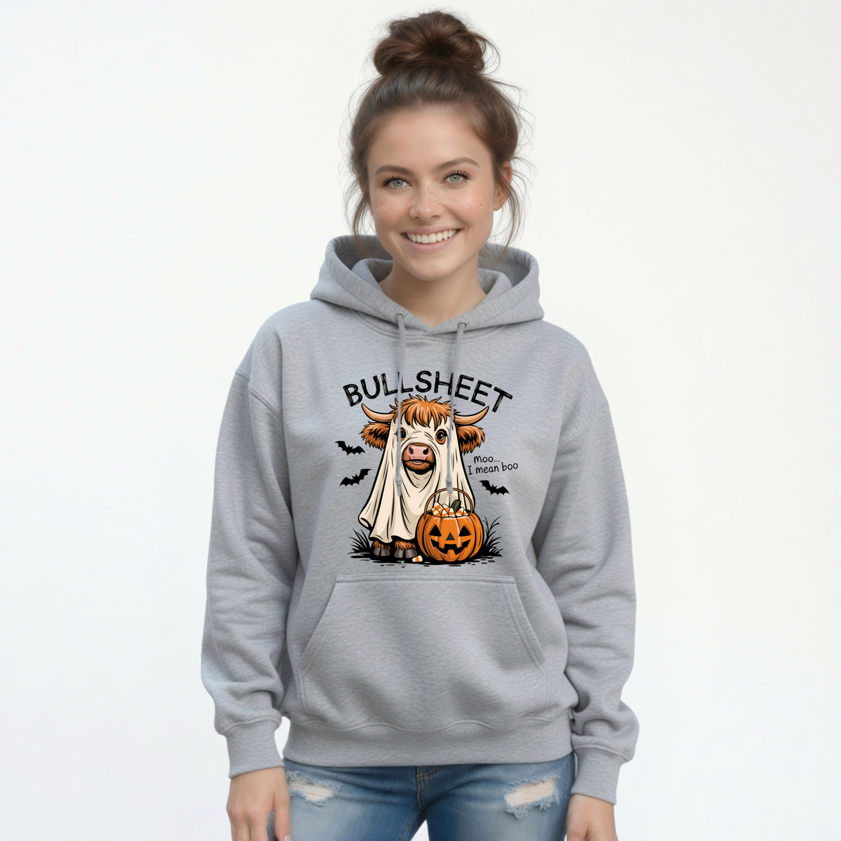 Smiling woman wearing a Sports Gray pullover hoodie featuring the Bullsheet Halloween Highland cow ghost and jack-o'-lantern design.