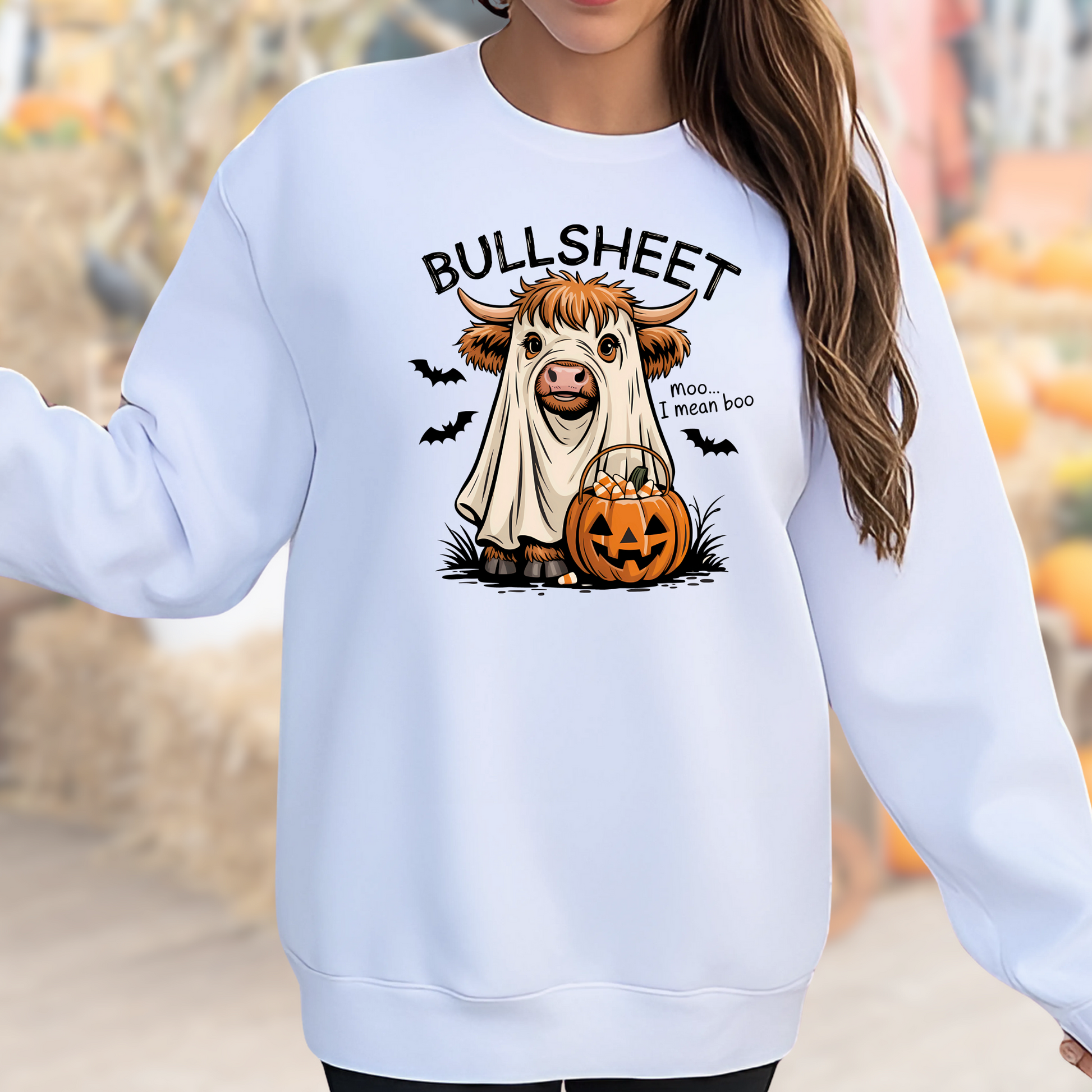 White sweatshirt with a Halloween-themed design of a cow in a sheet with a pumpkin and text, worn by a person.