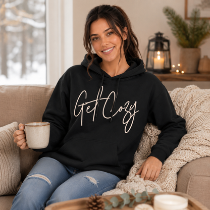 Woman wearing a black pullover hoodie with a minimalist Get Cozy script design while sitting on a couch and holding a mug in a cozy living room.