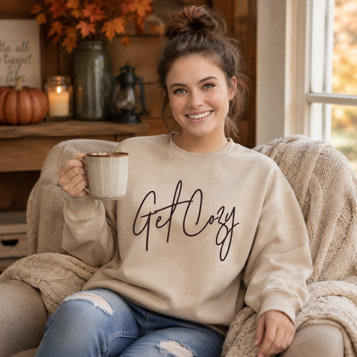 Woman wearing a sand crewneck sweatshirt with a minimalist Get Cozy script design while holding a mug in a warm, cozy living room.