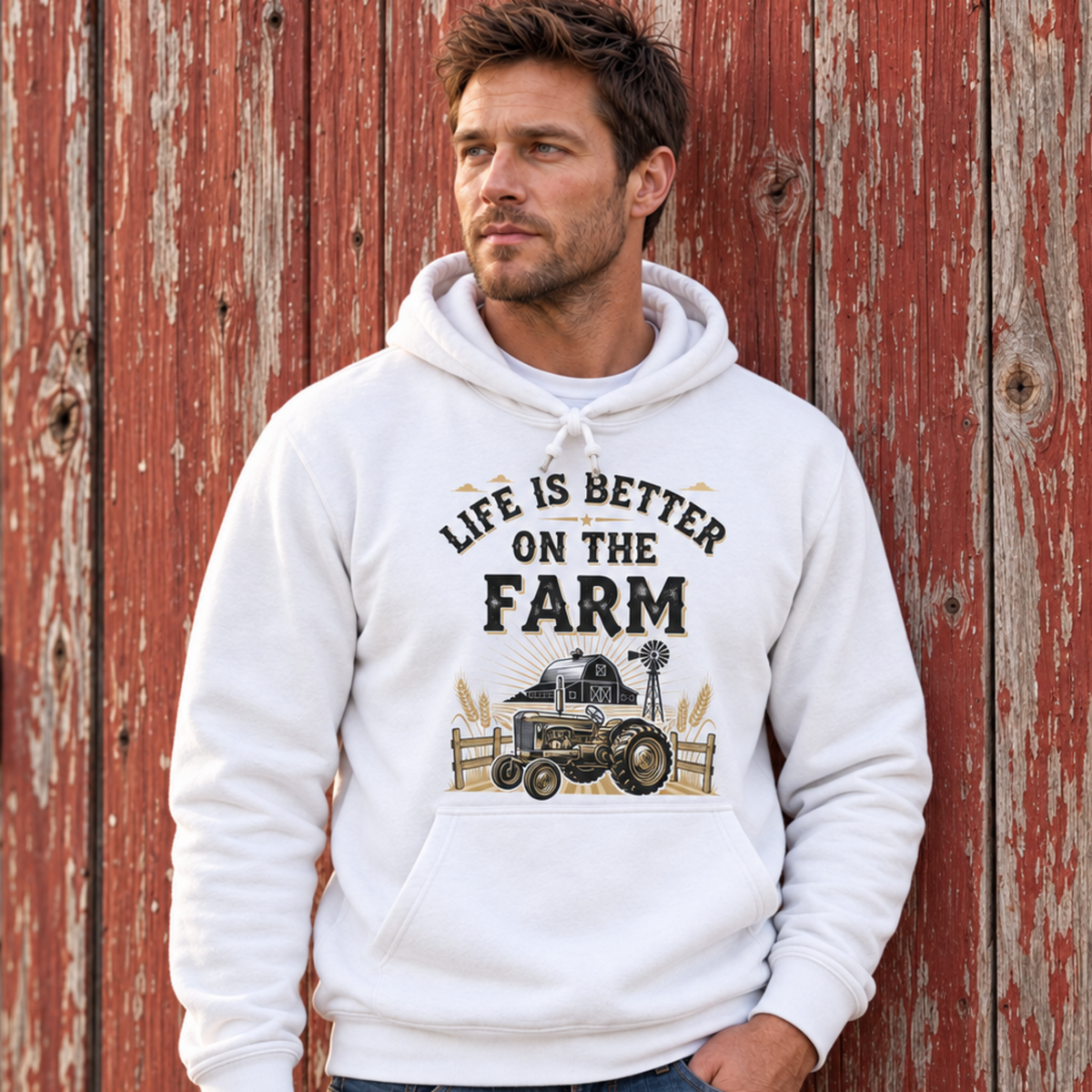 Man wearing a white Life Is Better on the Farm hoodie with tractor, barn, and windmill graphic against a weathered red barn wall.