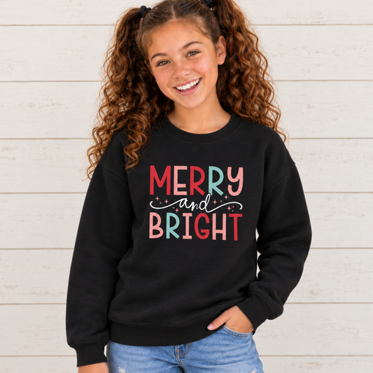 Girl wearing a black youth sweatshirt with colorful Merry and Bright Christmas design against a white wood wall