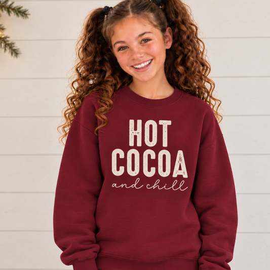 Youth girl wearing a maroon Hot Cocoa and Chill sweatshirt against a white shiplap wall