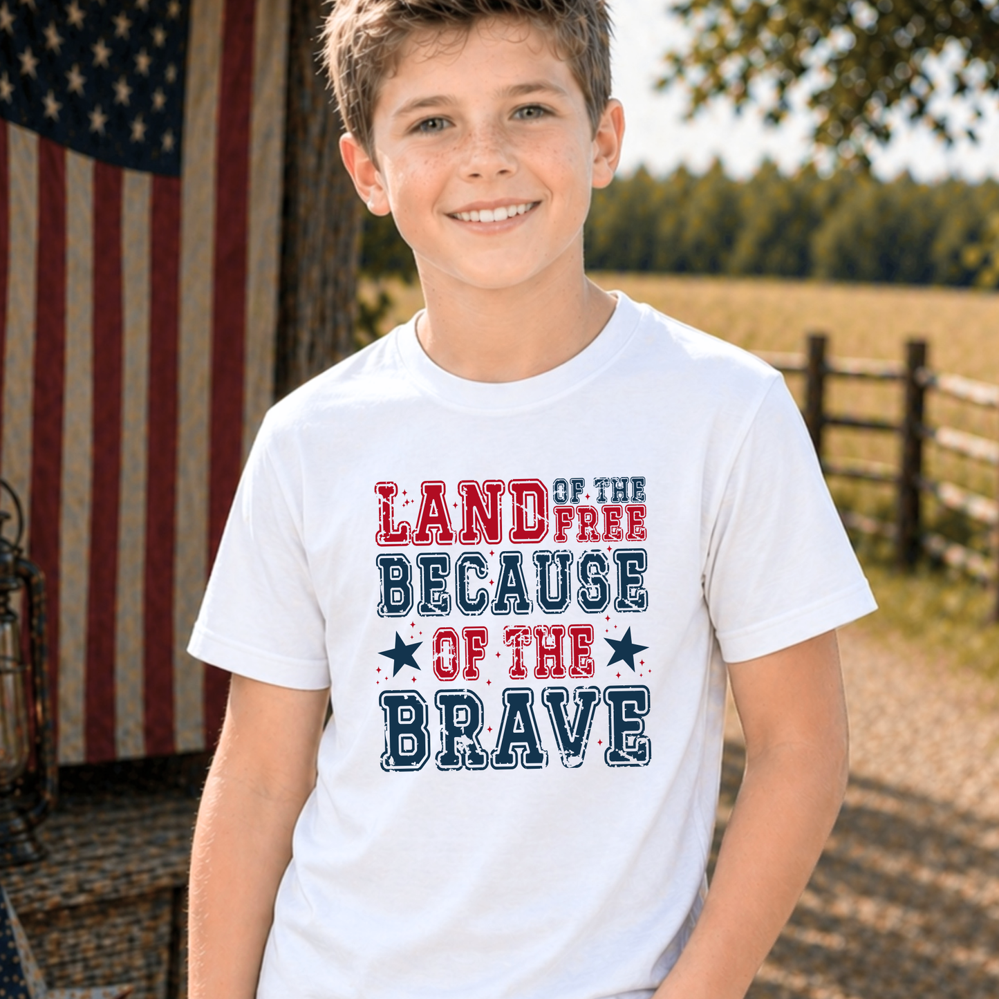 Child wearing a 'Land of the Free Because of the Brave' t-shirt with an American flag in the background.
