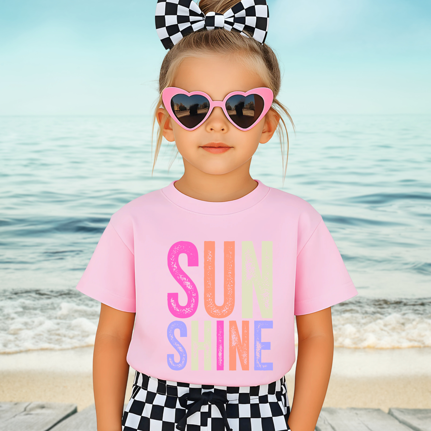 Child wearing a pink 'SUNSHINE' t-shirt with sunglasses and a checkered bow at the beach.