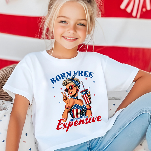 Child wearing a white t-shirt with 'Born Free But Now It's Expensive' graphic design in front of an American flag.