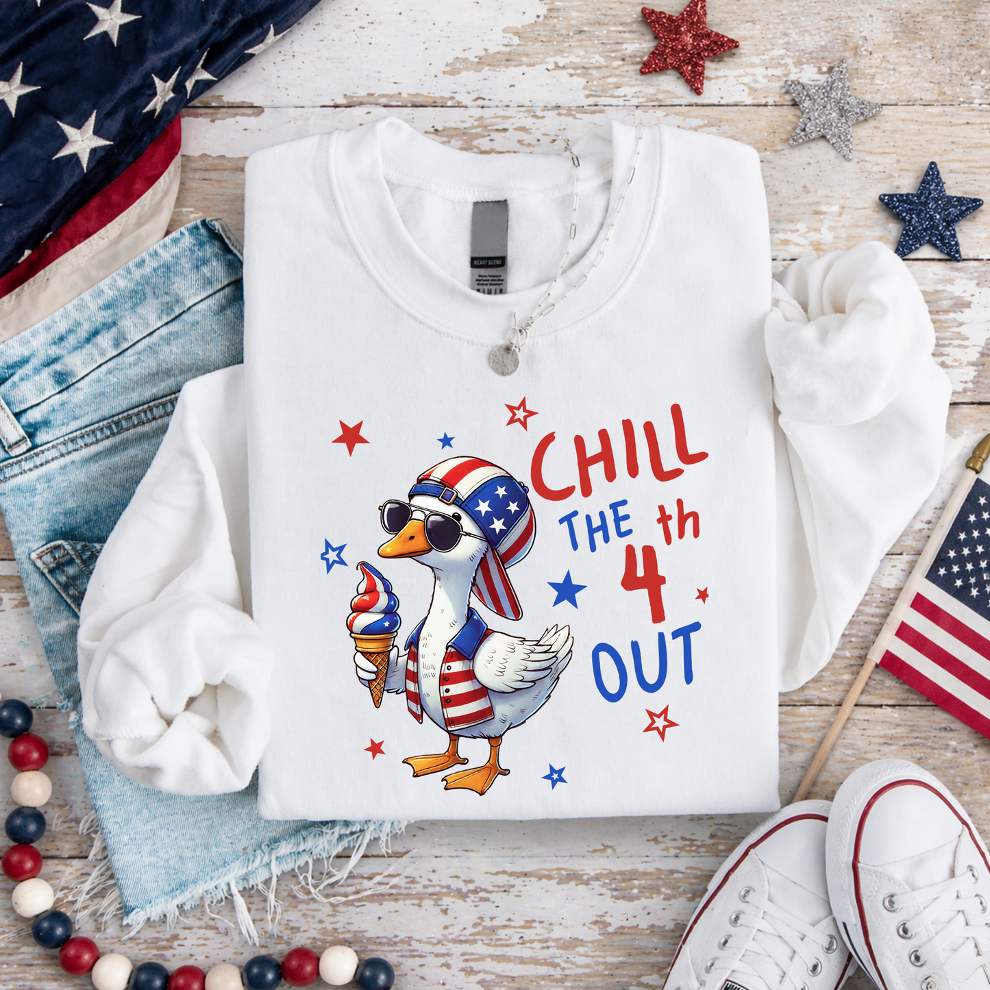 White sweatshirt with a graphic of a duck wearing an American flag hat and sunglasses, holding an ice cream cone, with text 'CHILL THE 4th OUT' on a wooden surface with American flags and stars.