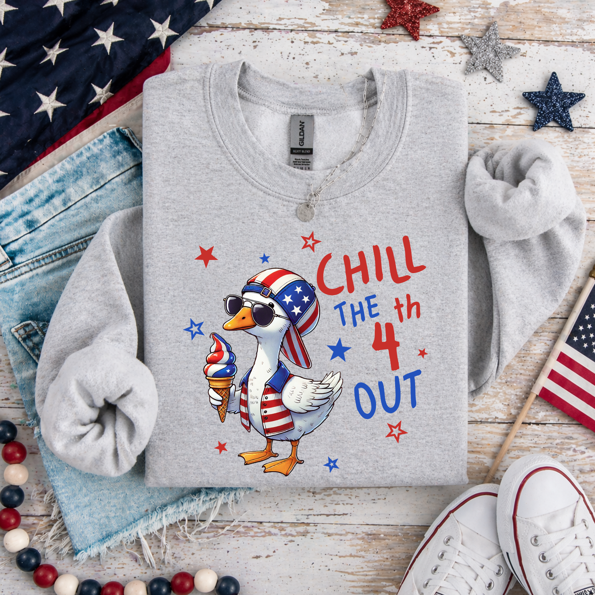 Gray sweatshirt with a duck wearing an American flag hat and ice cream, text 'Chill the 4th Out', on a wooden surface with American flags and jeans.