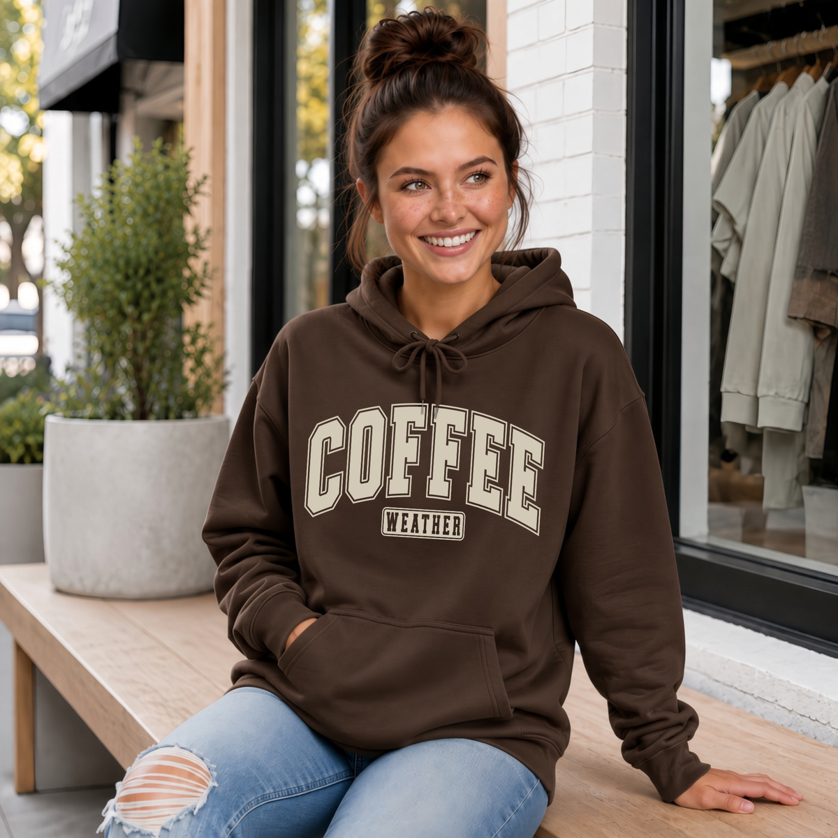 Woman sitting on a porch wearing a brown “Coffee Weather” crewneck sweatshirt and jeans.