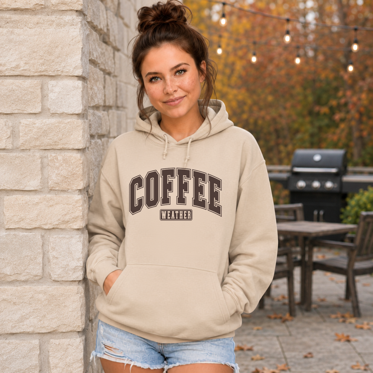 Woman outdoors wearing a sand-colored “Coffee Weather” hooded sweatshirt with denim shorts.