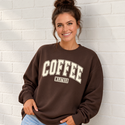 Smiling woman wearing a brown crewneck sweatshirt with a varsity-style “Coffee weather” graphic against a light brick wall